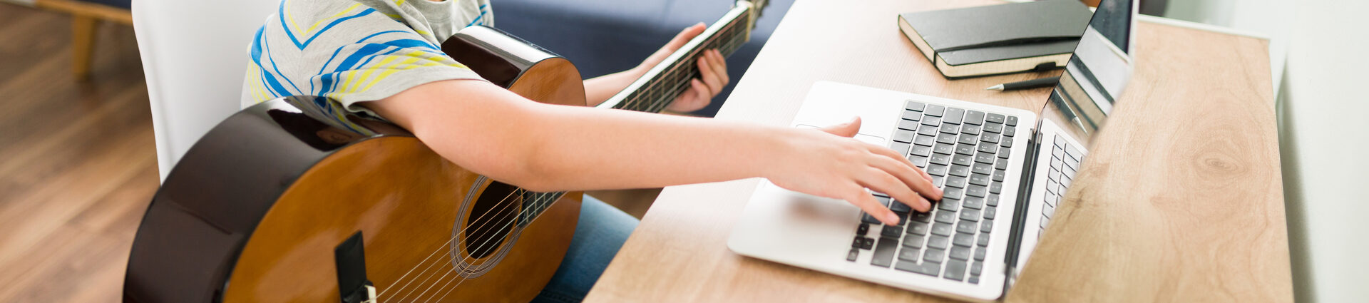 Ein Junge sitzt mit einer Gitarre vor einem Laptop und tippt etwas ein