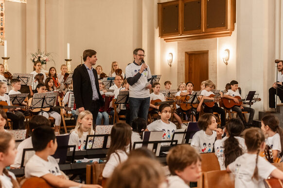 Ein Mann hält ein Mikrofon inmitten einer Schaar Kinder mit Musikinstrumenten