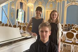 Zwei Frauen in dunklen Kleidern stehen hinter einem Mann, der ein schwarzes Hemd trägt und seitlich an einem weißen Klavier sitzt.